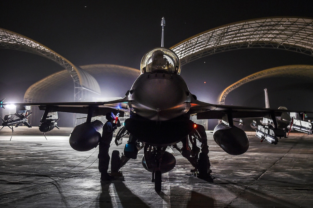 Airmen work on a jet sitting on a flightline at night, with curved hangar roofs illuminated in the background.