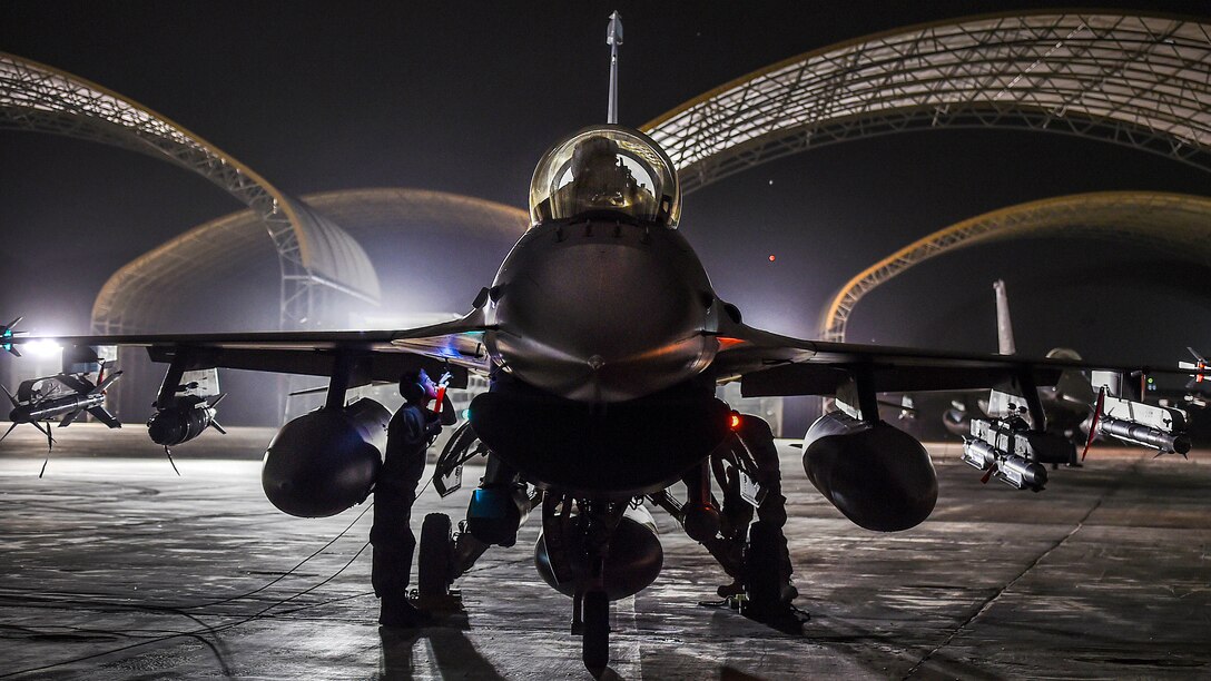 Airmen work on a jet sitting on a flightline at night, with curved hangar roofs illuminated in the background.