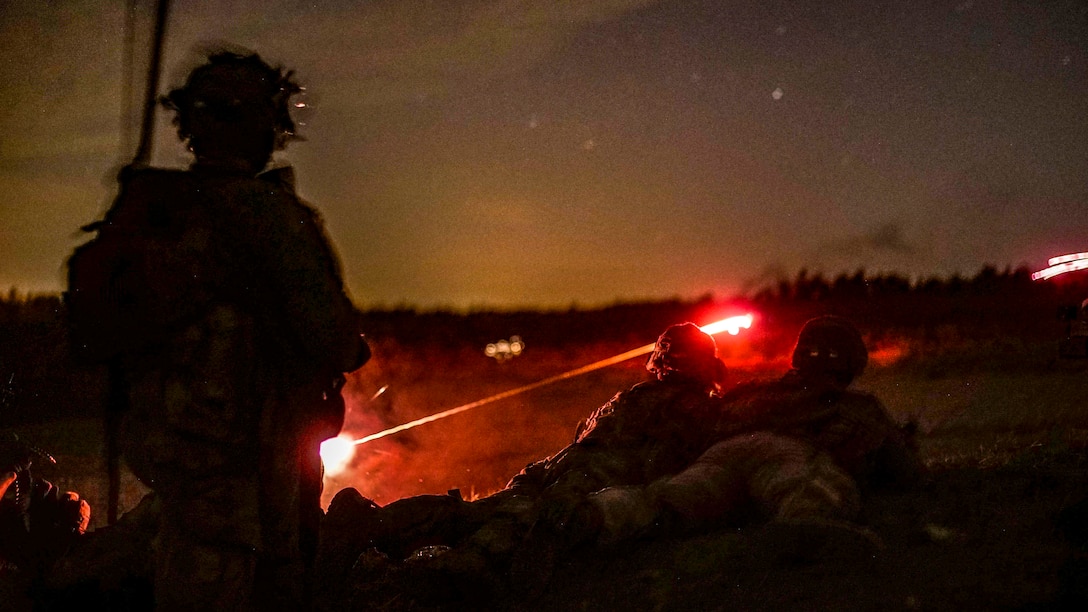 Red light shoots down a dark range as soldiers, shown in silhouette, fire weapons.