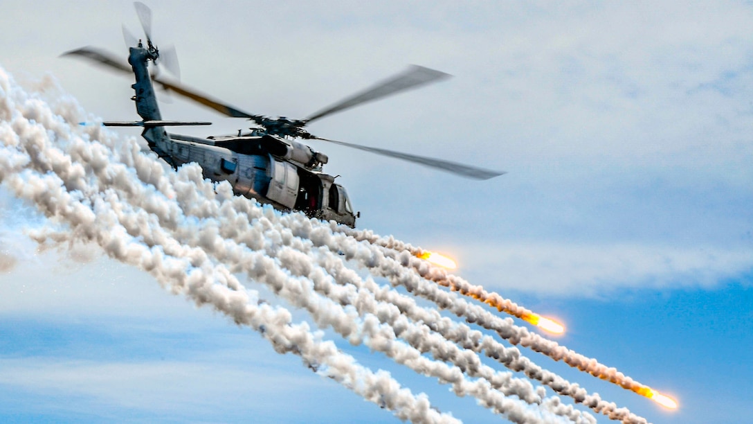 A helicopter flies in front of several cloud trails from flares it is firing in a blue sky.