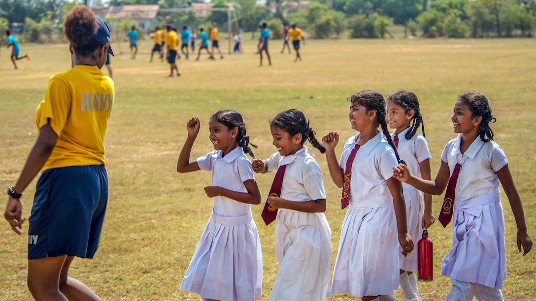 Five girls in matching white school uniforms laugh as they move in a line behind a sailor.