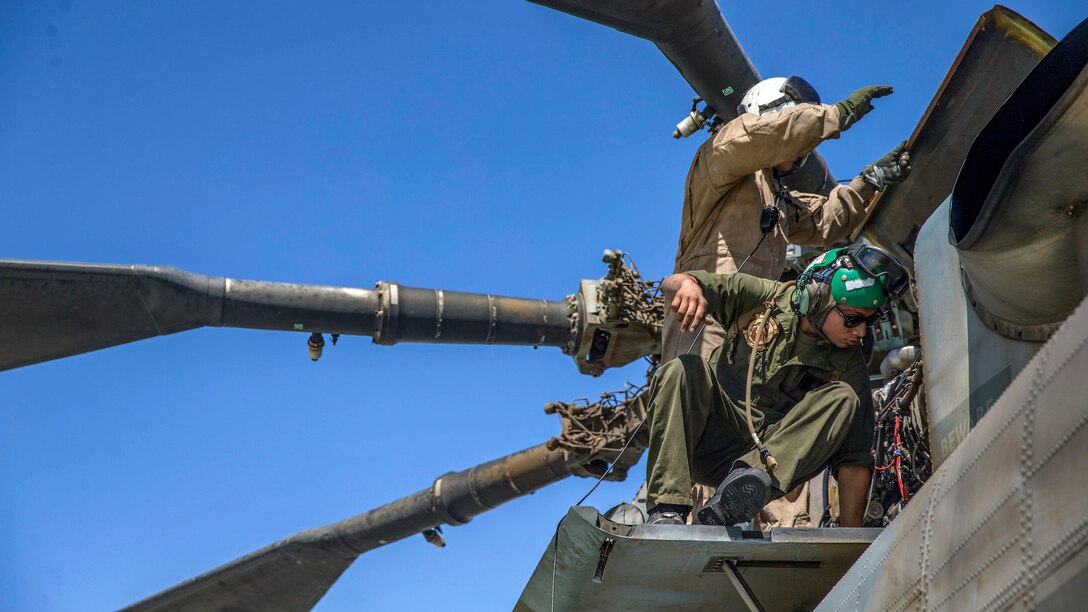 Two Marines in helmets inspect the top of a helicopter, as the rotor blades fan out against a blue sky.