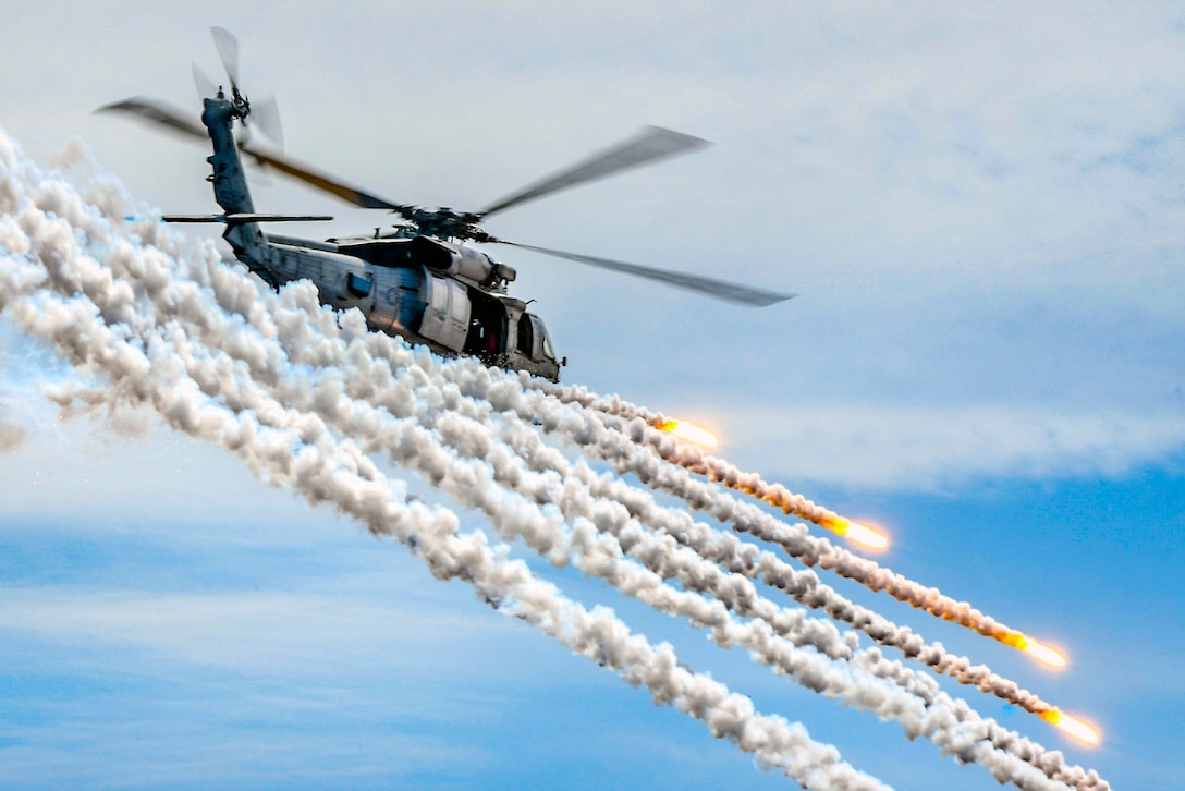 A helicopter flies in front of several cloud trails from flares it is firing in a blue sky.