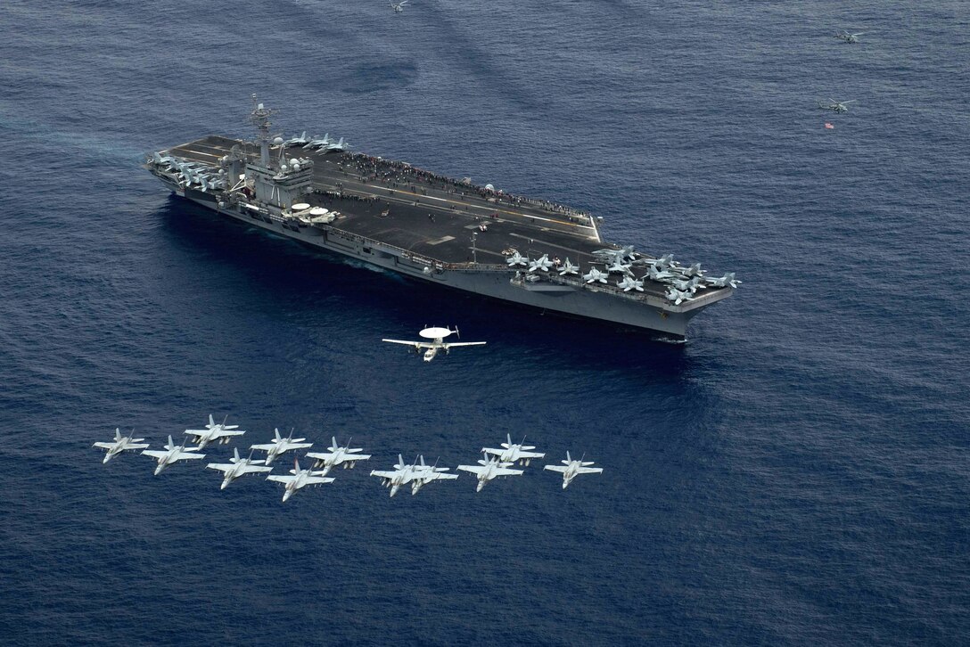 Aircraft fly in formation above the aircraft carrier USS Theodore Roosevelt.