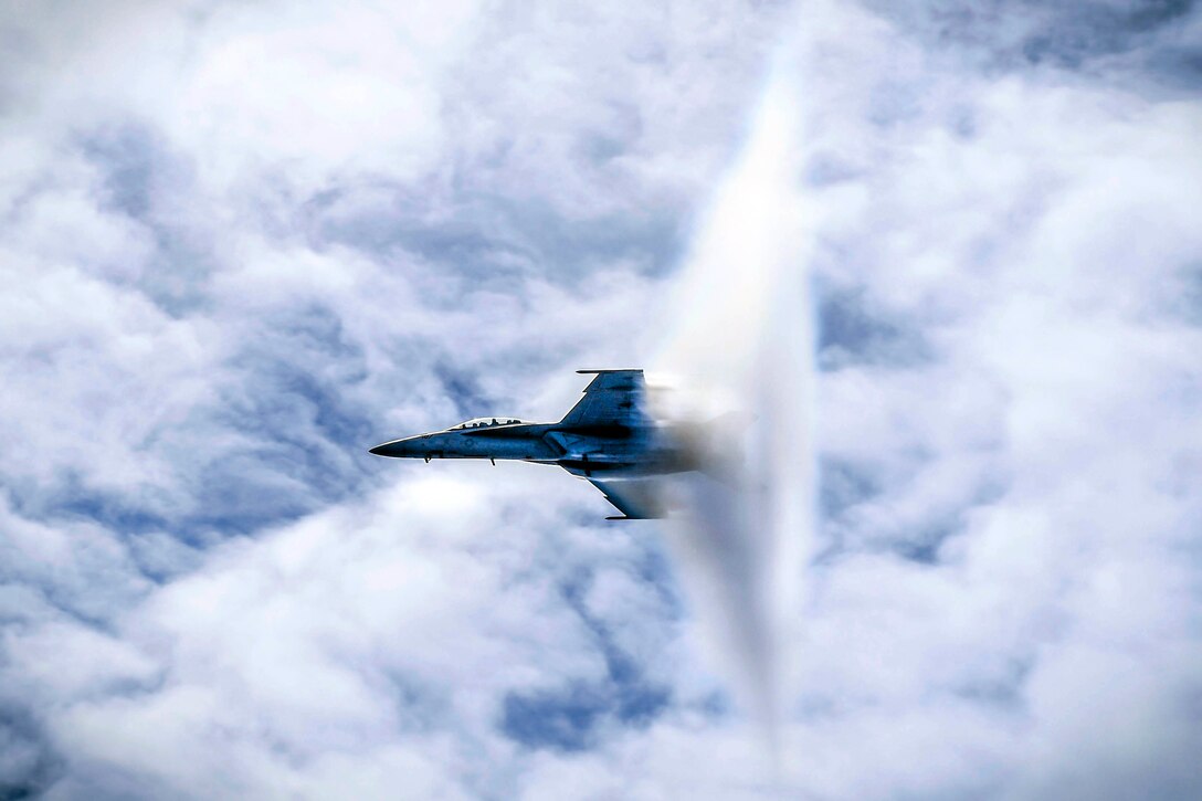 A funnel cloud forms around the tail of a jet flying in blue cloudy sky.