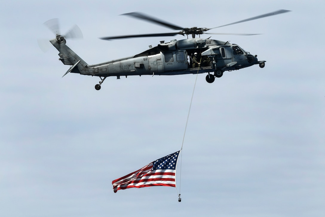 An MH-60S Seahawk helicopter flies the U.S. flag alongside the aircraft carrier USS Theodore Roosevelt.