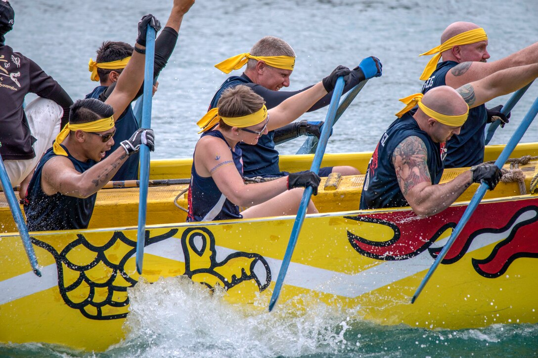 Navy personnel wearing yellow bandannas, row in a yellow boat with dragon-themed decoration on the side.