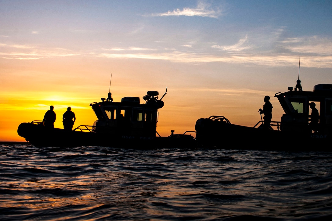 Airmen, shown in silhouette, look out from two small boats in water, against an orange and blue sky.