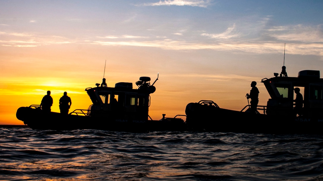 Airmen, shown in silhouette, look out from two small boats in water, against an orange and blue sky.