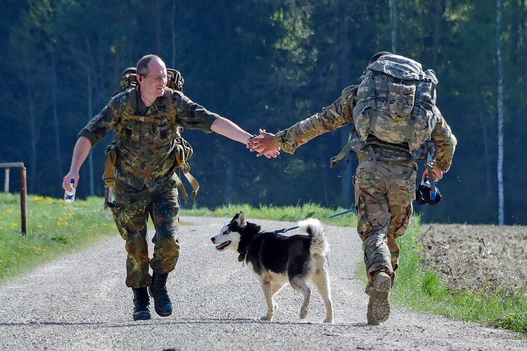 A U.S. soldier with a backpack, shown from behind, claps hands with a German soldier walking in the other direction on a gravel road.