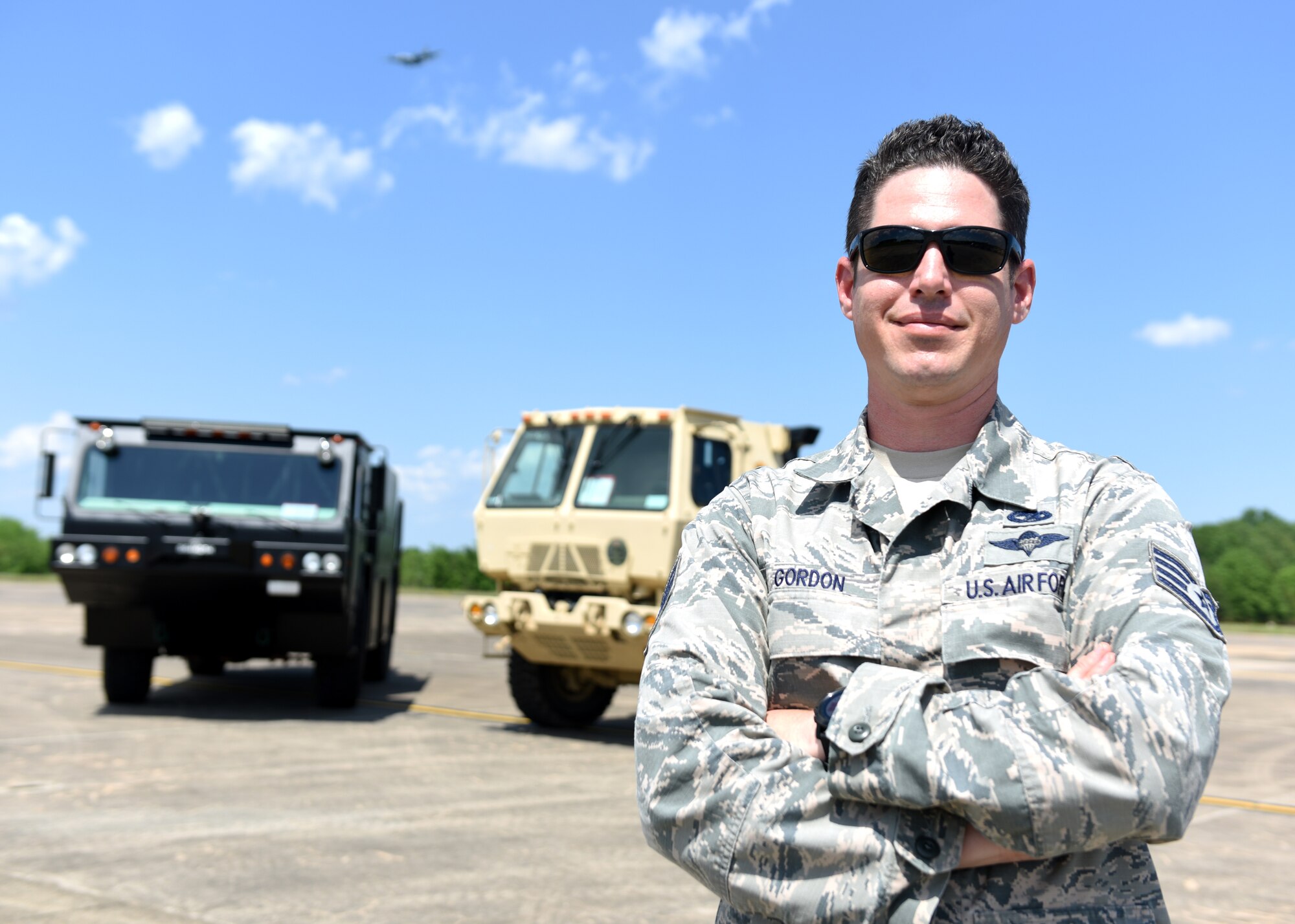 A male in uniform stands on a bright flightline in front of two vehicles.