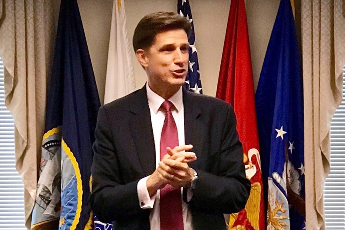 Dana S. Deasy stands and speaks in front of flags in a room.