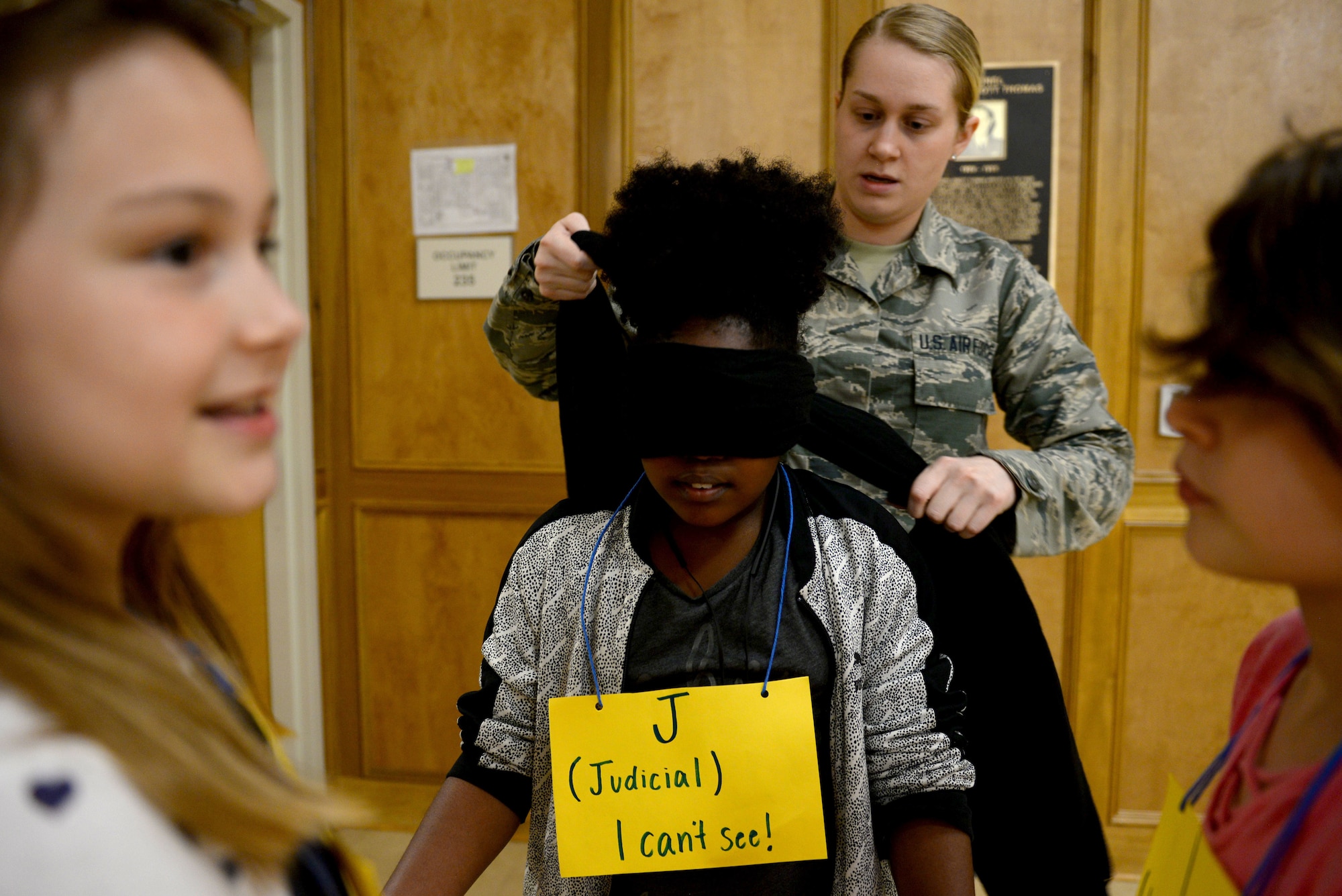 A young girl gets a black scarf tied around her eyes by a woman in the Airman Battle Uniform and two girls smile at the edge of the photo.