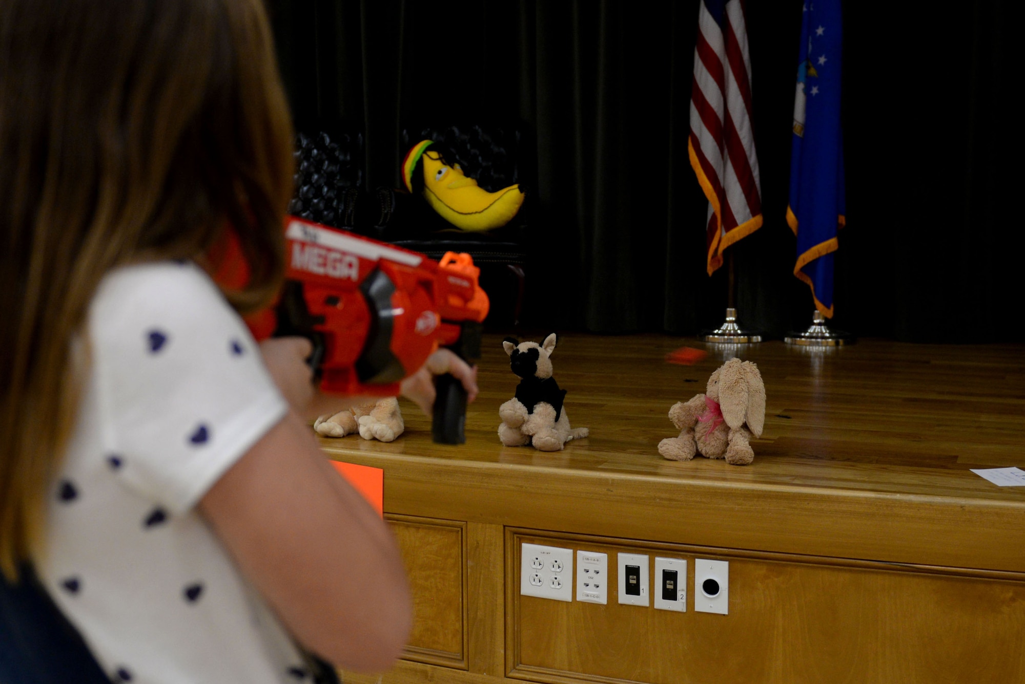 A young girl wearing a white shirt with blue hearts points a dart gun at stuffed animals on a stage.