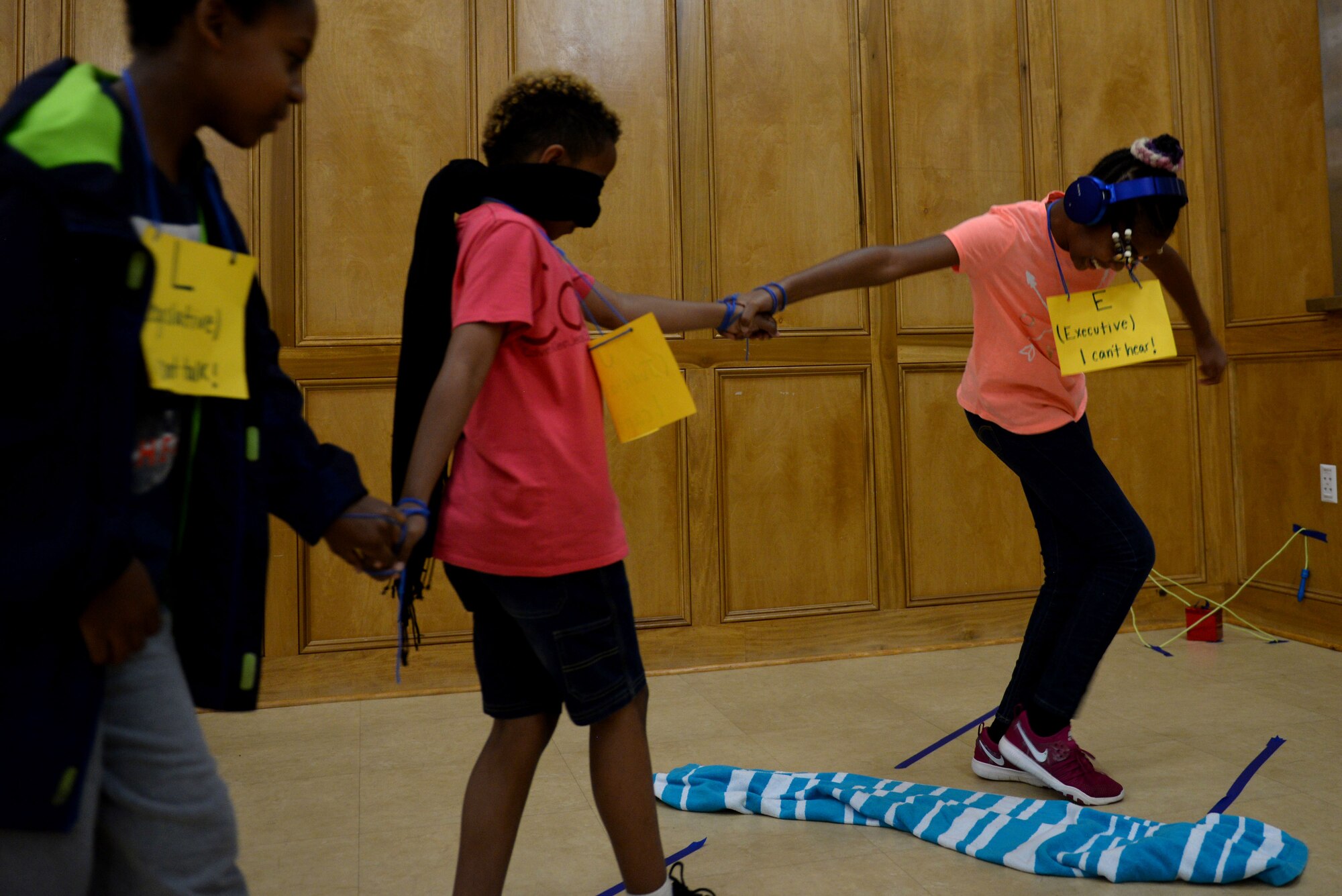 A young girl wearing an orange top and black pants leaps over a blue and white striped towel while holding hands with a boy in a red shirt and black shirt who's holding hands with another young boy wearing a black an green hoodie and blue jeans.