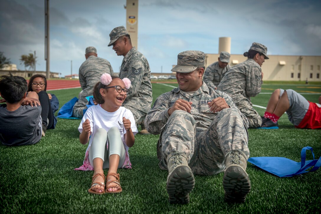A girl and an airman look at each other and laugh while finishing a situp on a field outside.