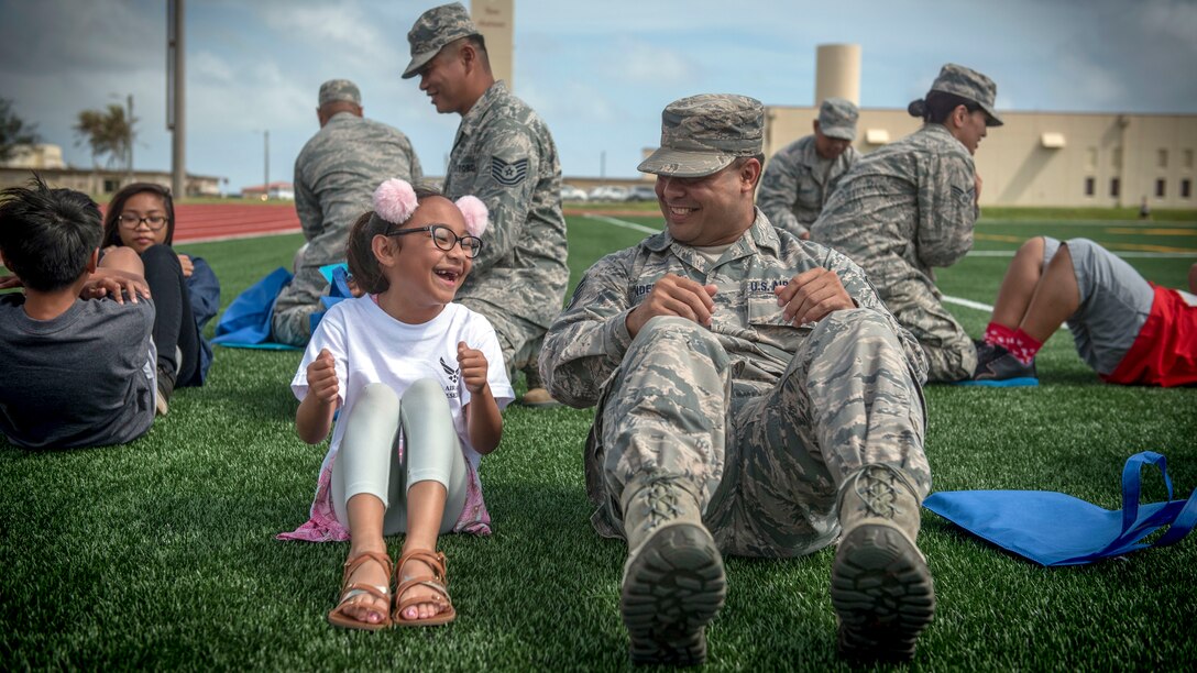 A girl and an airman look at each other and laugh while finishing a situp on a field outside.