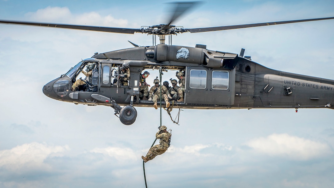 A soldier hangs from a rope from a helicopter, where fellow soldiers are sitting by an open door.
