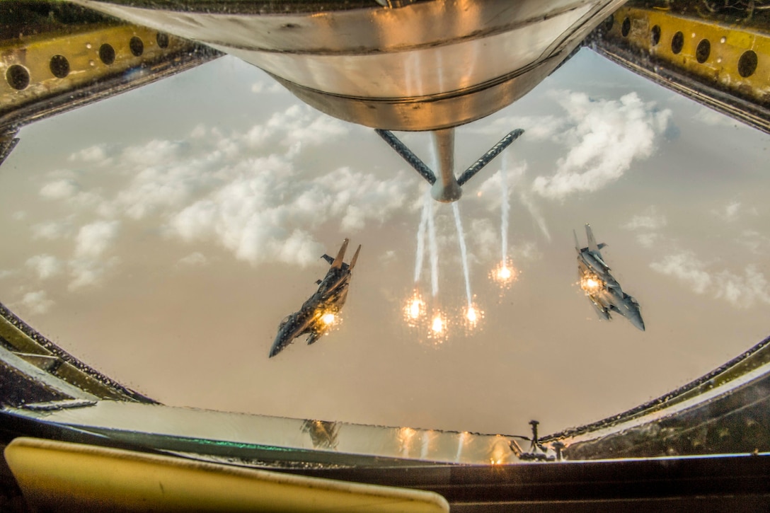 Two jets, framed by an opening in an aircraft in front of them, fire flares while in flight.