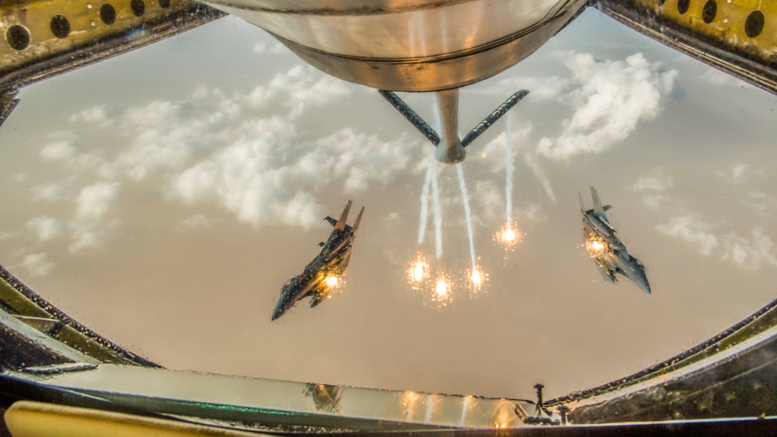 Two jets, framed by an opening in an aircraft in front of them, fire flares while in flight.