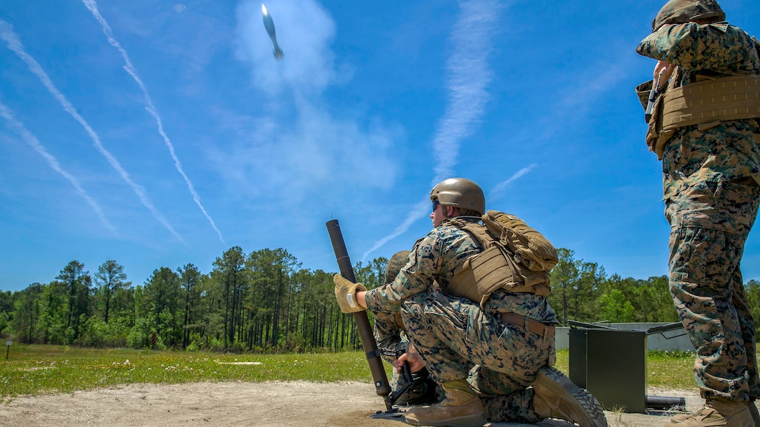 A Marine kneels and fires a mortar system, sending a projectile into blue sky, as a fellow Marine stands by.