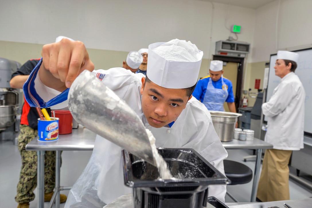 A sailor wearing white chef's clothing pours flour from a scoop into a plastic container.