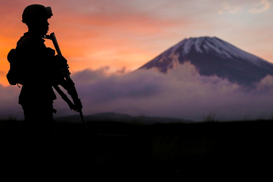 A Marine, shown in silhouette, stands with a weapon against an orange and purple cloudy sky, with Mount Fuji in the background.