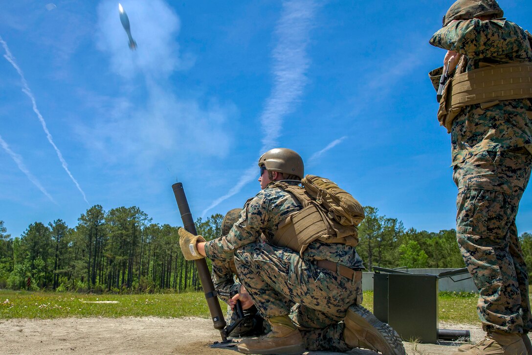 A Marine kneels and fires a mortar system, sending a projectile into blue sky, as a fellow Marine stands by.