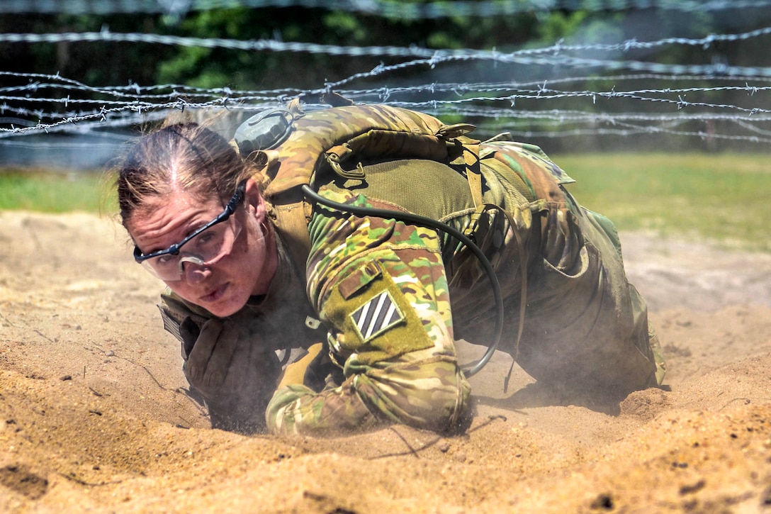 A soldier crawls in sand under barbed wire.