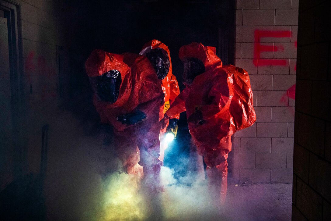 Three service members in orange protective garb stand together in a smoky room.