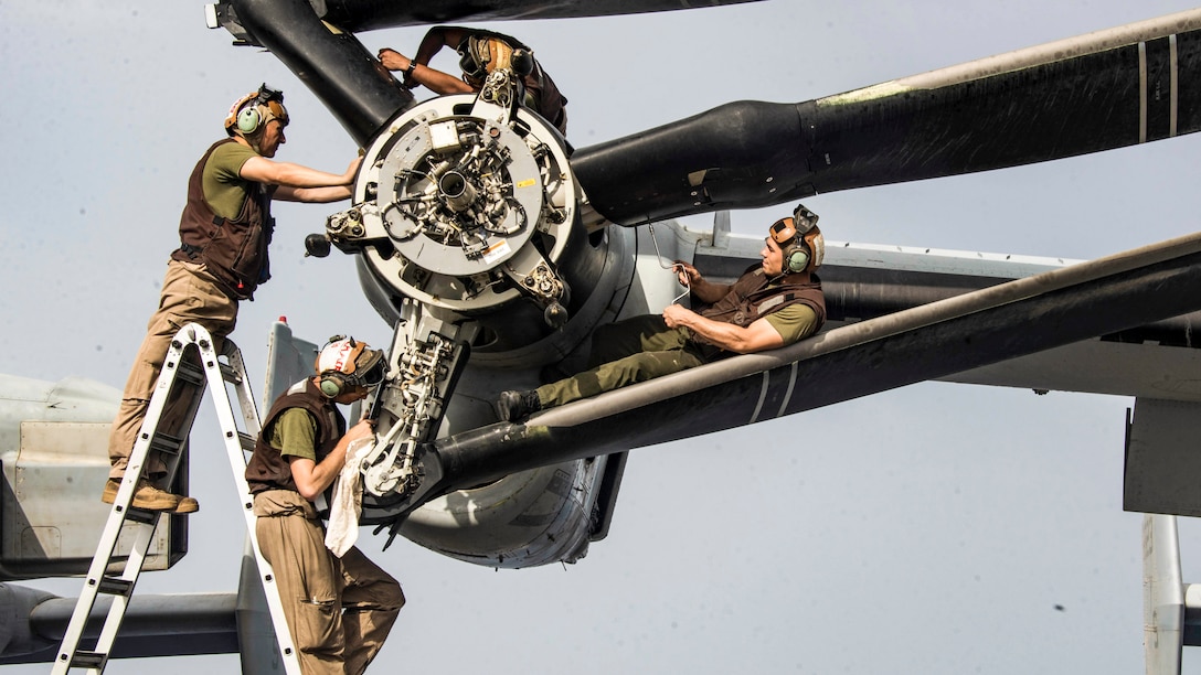 Two Marines stand on a ladder and conduct maintenance on an aircraft, and two lie on aircraft wings and do the same.