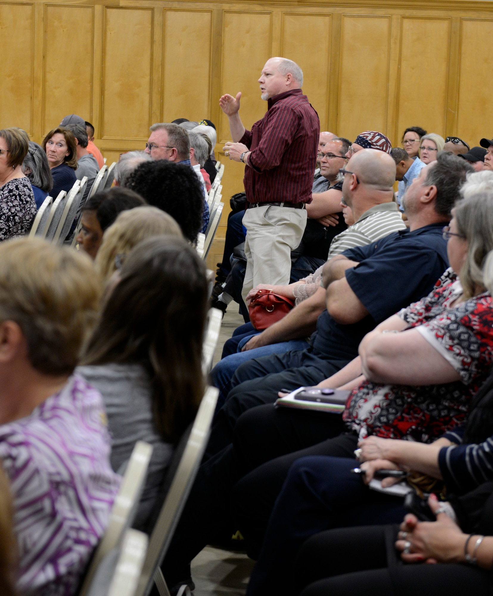 An older white male stands in the middle of a seated crowd to ask a question.