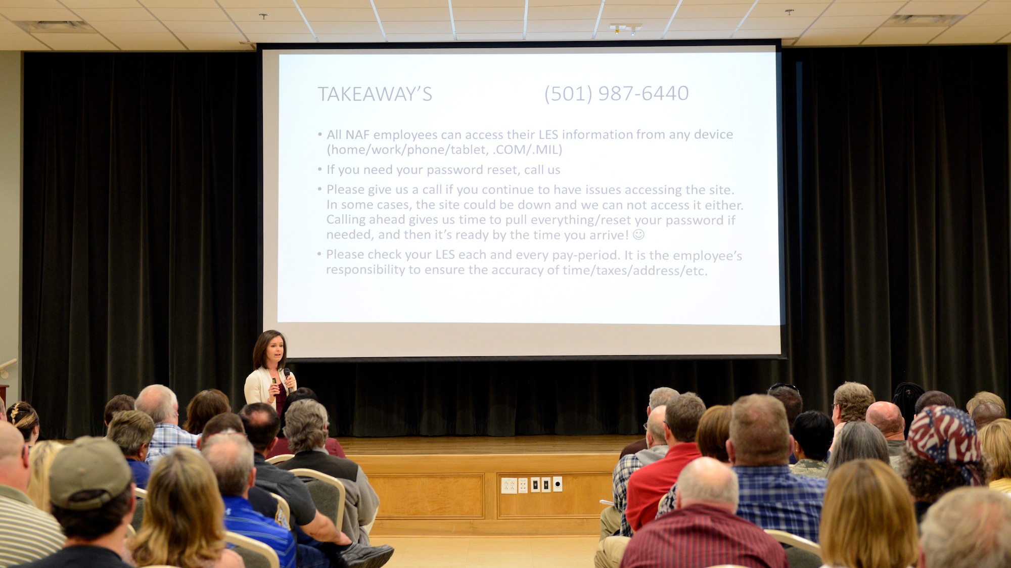 Young white adult female stands in front of seated crowd talking about the slide projected on the screen behind her.
