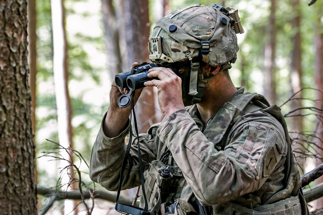 A soldier looks through binoculars for opposing force soldiers.
