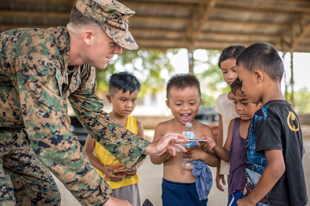 A Marine hands a sticker to a little boy, as others gather around smiling.
