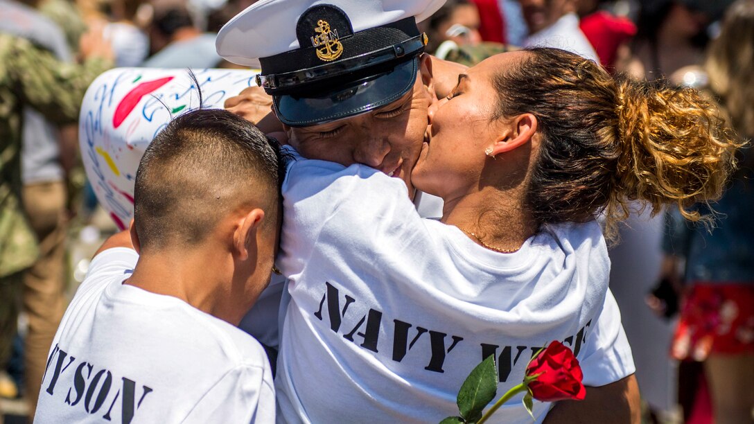 A sailor hug his wife, who kisses his cheek, and child, who buries his head into the embrace.