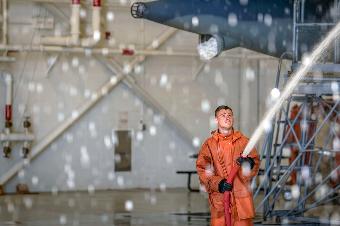 Droplets of water fill the frame as an airman in an orange raincoat sprays an aircraft in a hangar.