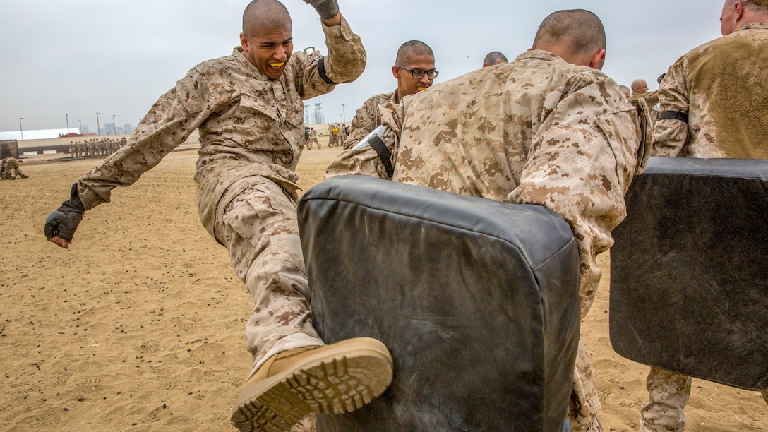 A recruit wearing a mouth guard kicks into a black cushion held by another recruit on sandy terrain.
