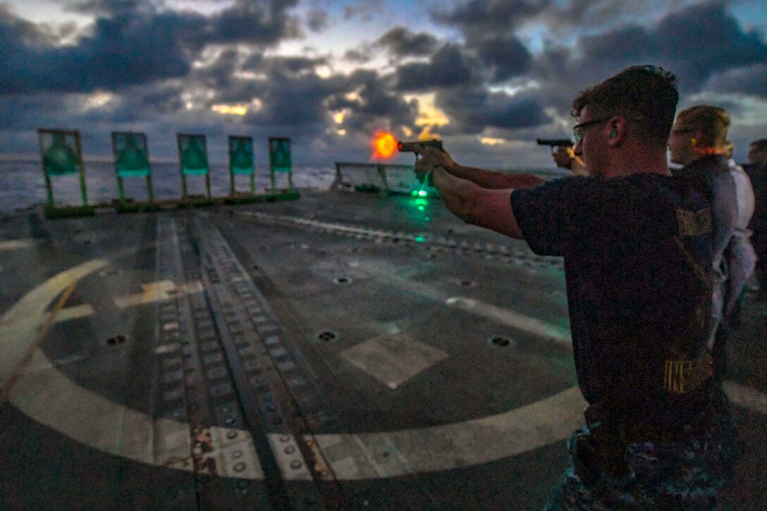 An orange flash emits from a pistol as a sailor fires at a target on a ship's flight deck.