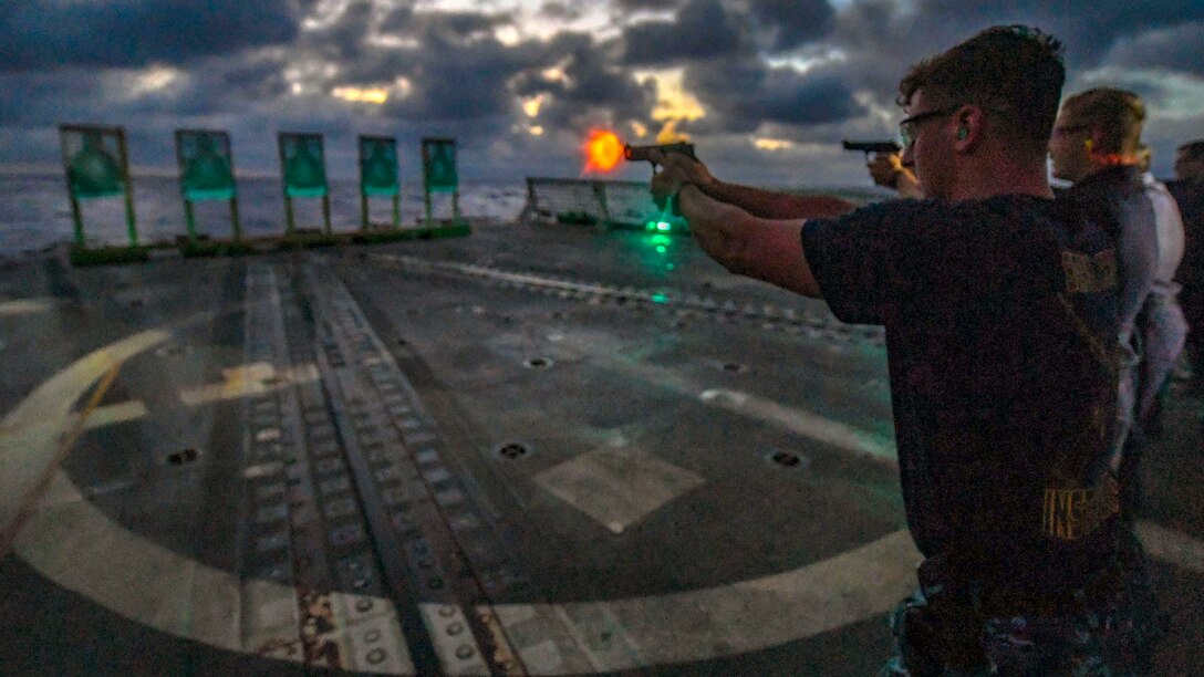 An orange flash emits from a pistol as a sailor fires at a target on a ship's flight deck.