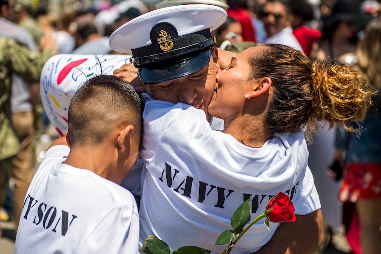 A sailor hug his wife, who kisses his cheek, and child, who buries his head into the embrace.