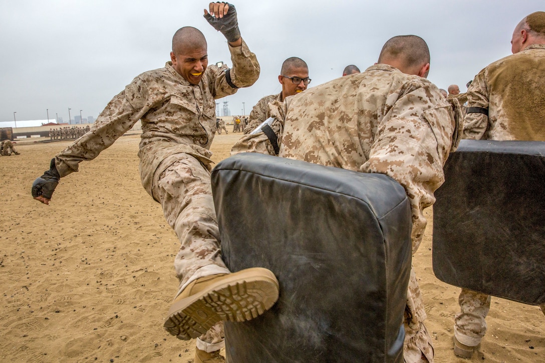 A recruit wearing a mouth guard kicks into a black cushion held by another recruit on sandy terrain.