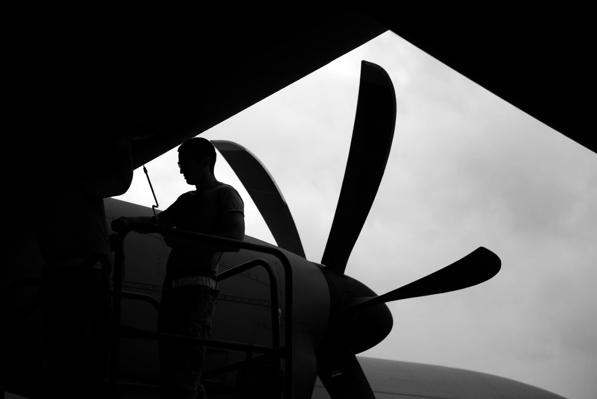 A main who is silhouetted takes a took to the wing of a C-130J with a propeller in the background.