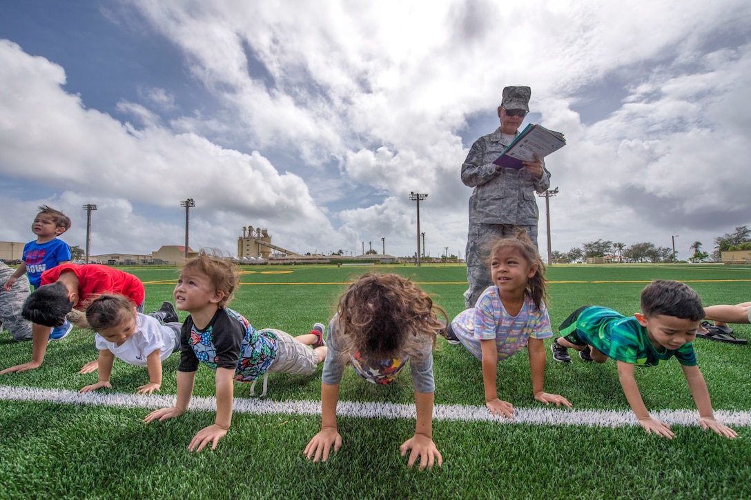 Children do pushups in a row on an athletic field as an airman stands over them watching.
