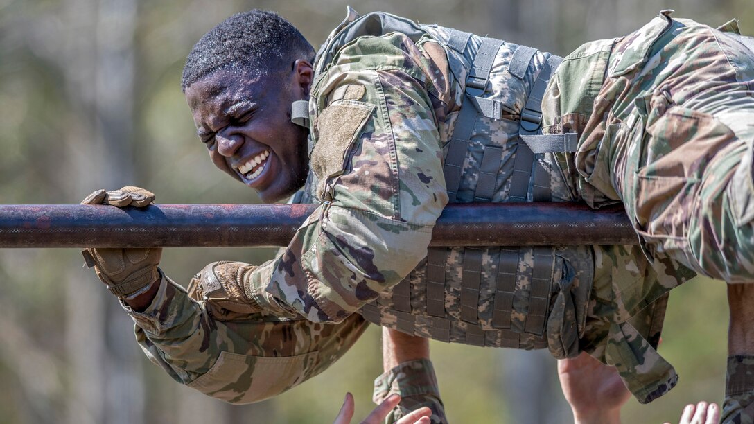 A soldier winces while holding onto a horizontal metal bar, as fellow troops' hands reach up to support him.