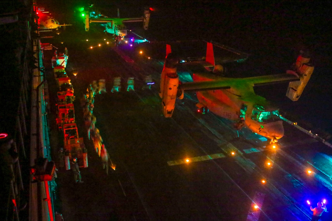 Marines walk in a curved line to board an Osprey aircraft at night on a flight deck, illuminated by colored lights.