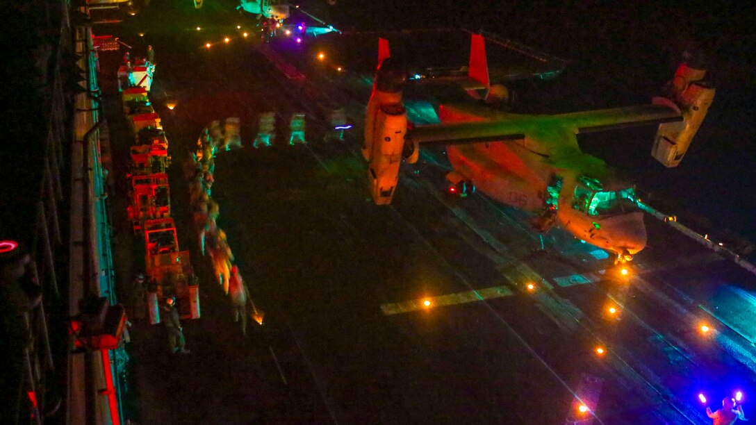 Marines walk in a curved line to board an Osprey aircraft at night on a flight deck, illuminated by colored lights.