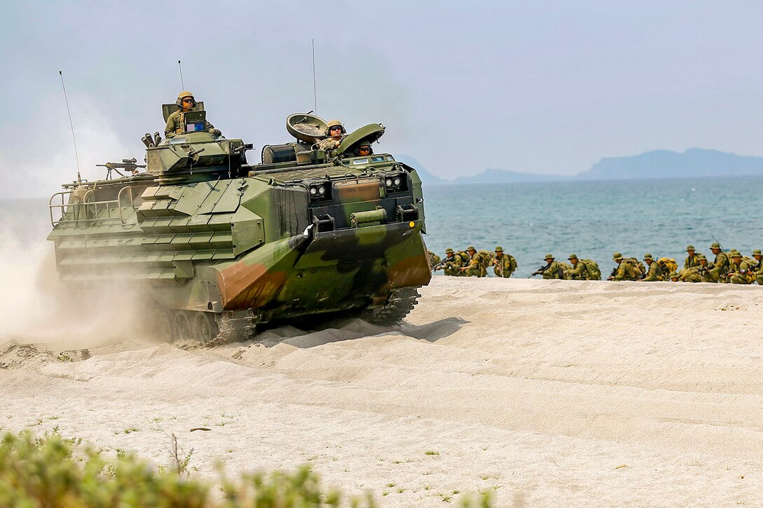An amphibious vehicle kicks up sand while driving on the beach, as troops crouch in a line nearby.