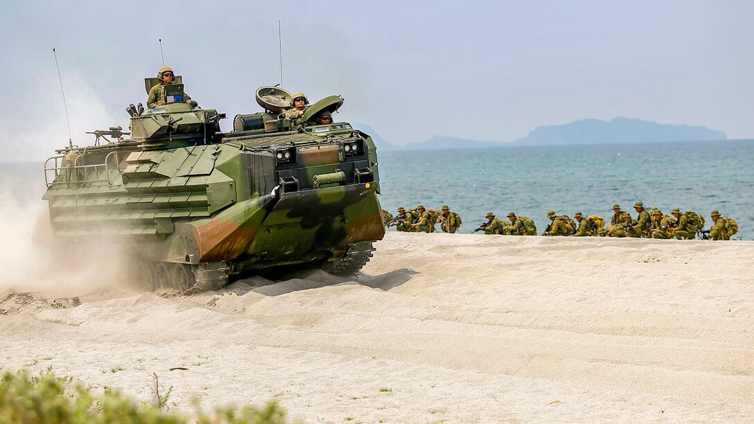 An amphibious vehicle kicks up sand while driving on the beach, as troops crouch in a line nearby.