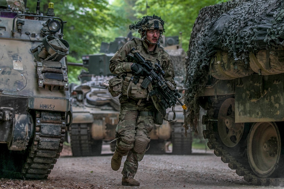 A soldier flanked by tactical vehicles jogs with a weapon on a roadway.